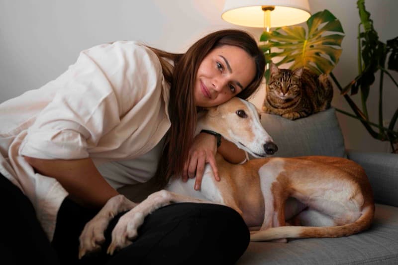 Mujer abrazando a su galgo en el sofá de casa con un gato al lado